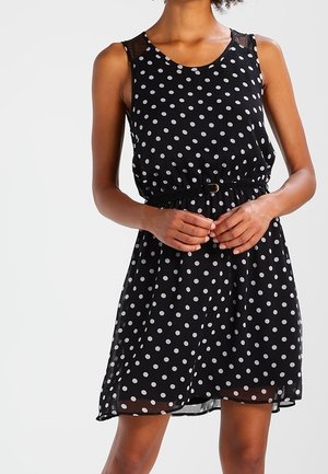 Czarna sukienka bez rękawów w białe kropki, z paskiem w talii, do kolan, noszona przez stojącą osobę ze złożonymi przed sobą rękami.