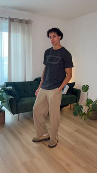 Νεαρό άτομο με σγουρά μαλλιά στέκεται σε ένα καθιστικό φορώντας μαύρο μπλουζάκι, μπεζ παντελόνι με φαρδιά πόδια και ταμπά μοκασίνια κοντά σε μια πράσινη καναπέ και φυτά.