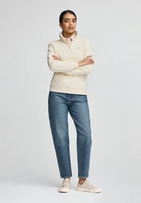 Felpa a mezza zip color beige chiaro, jeans blu e sneakers beige. La felpa presenta un piccolo logo sul petto e polsini a coste.
