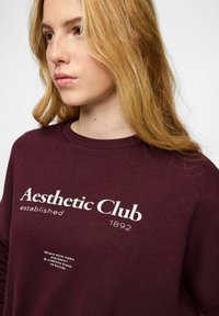 Młoda kobieta o długich blond włosach, ubrana w ciemno bordową bluzę z napisem "Aesthetic Club established 1892" na przodzie.