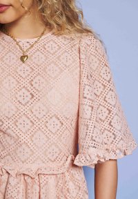 Mulher usando um vestido de renda floral rosa com bordas em babados e um colar com pingente em forma de coração dourado, contra um fundo azul.