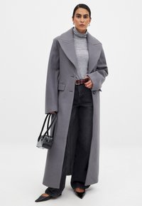 Lång grå kappa med breda slag, bärs över en ljusgrå polotröja, kombinerat med högmidjade svarta jeans och spetsiga svarta skor. Liten svart handväska.