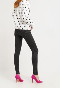 Kobieta w białej koszuli z długim rękawem w czarne kropki, czarnych obcisłych jeansach oraz jasnoróżowych butach na wysokim obcasie, stojąca bokiem.