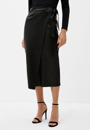 Jupe portefeuille en similicuir noir midi avec un noeud sur le côté, portée avec des sandales noires à talons hauts et bride à la cheville, ainsi qu'un haut à manches longues.