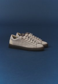 Paire de baskets basses en cuir beige avec des lacets blancs et un détail perforé sur le côté sur un fond bleu foncé.