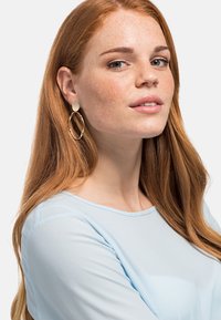 Guldfarvede øreringe med en oval stud og en forlænget, snoet hoops, sat mod en lys blå bluse. Glat metaloverflade.