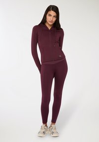 Giacca atletica bordeaux con cappuccio e chiusura a zip, abbinata a leggings coordinati. Tessuto resistente e con dettagli del logo.