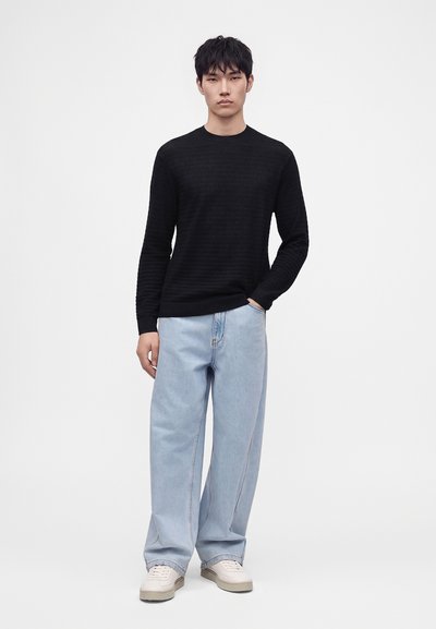 Jeune homme debout, une main dans la poche, portant un pull noir à manches longues, un jean large bleu clair et des baskets blanches sur fond blanc.