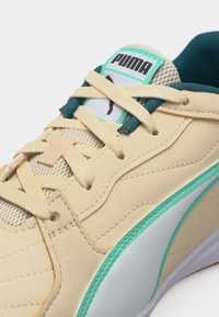 Μπεζ αθλητικό παπούτσι Puma με τιρκουάζ λεπτομέρειες, λευκή σόλα και μπεζ κορδόνια δεμένα πάνω σε δίχτυ που αναδεικνύει το λογότυπο Puma.