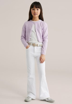 Dívka v lila svetříku, bílém tričku, bílých zvonových kalhotách s páskem a stříbrných teniskách, stojící s jednou rukou v kapse.