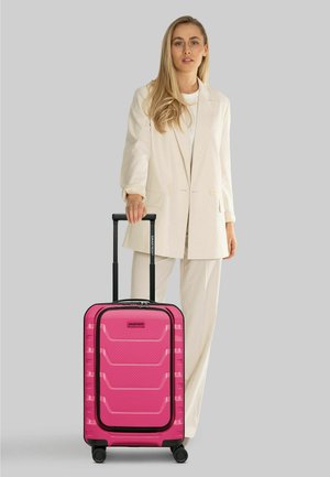 Mujer con traje beige de pie junto a una maleta rígida rosa con asa negra extendida sobre fondo gris.