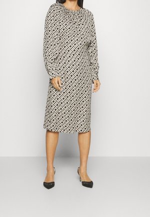 Kobieta w sukience o długości do kolan, z długimi rękawami, w czarno-biały geometryczny wzór, ubrana w czarne szpilki z ostrym noskiem, na jednolitym tle.