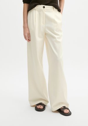 Personne portant un pantalon crème à jambes larges avec des plis à l'avant, un haut texturé foncé et des sandales noires à brides sur fond blanc.