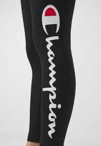 Czarne legginsy wykonane z elastycznego materiału, z wyraźnym białym logo "Champion" i czerwonym akcentem na lewym udzie.