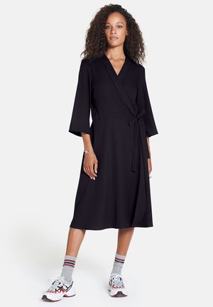 Schwarzes Wickelkleid mit Dreiviertelärmeln, Bindegürtel und glatter Textur. Kombiniert mit weißen Sneakers und grauen Socken mit roten Streifen.