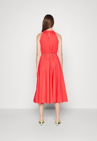 Femme aux longs cheveux bruns portant une robe midi rouge sans manches cintrée par une ceinture en chaîne et des sandales à talons jaunes, se tenant devant un mur blanc.