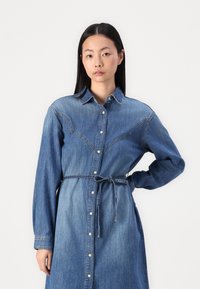 Jeans obleka v srednje modri barvi, z gumbom na sprednji strani, dolgimi rokavi, pasom in okrasnim šivanjem ob yoku.
