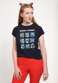 Mörkblå t-shirt med nio tecknade panels av Kalle Anka med olika uttryck, med vit text som säger "KALLE ANKAS HUMÖR". Röda byxor används.