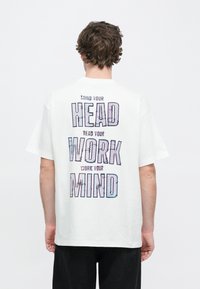 Biały bawełniany t-shirt z dużym, wielokolorowym nadrukiem z tyłu o treści „UWAŻAJ NA GŁOWĘ, ĆWICZ UMYSŁ”. Krótkie rękawy.