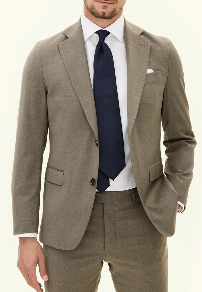Lichtbruin op maat gemaakt pak met een textuurafwerking, enkelvoudige blazer en gelapte revers. Gekozen met een marineblauwe stropdas en een wit shirt.