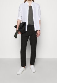 Camicetta di lino bianca, t-shirt grigia a V, pantaloni neri con cordoncino e sneaker bianche. Tiene una borsa verde scuro. Outfit semplice e informale.