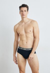 Schwarze Slips mit einem grauen Nike-Logo-Bund, ausgestattet mit einem figurbetonten Schnitt und glattem Stoff. Das Model steht mit neutralem Ausdruck.