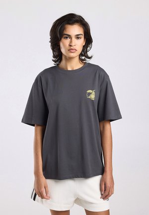 Vrouw die een losse donkergrijze T-shirt draagt met een gele fruitafbeelding op de borst en witte shorts met zwarte strepen aan de zijkanten.