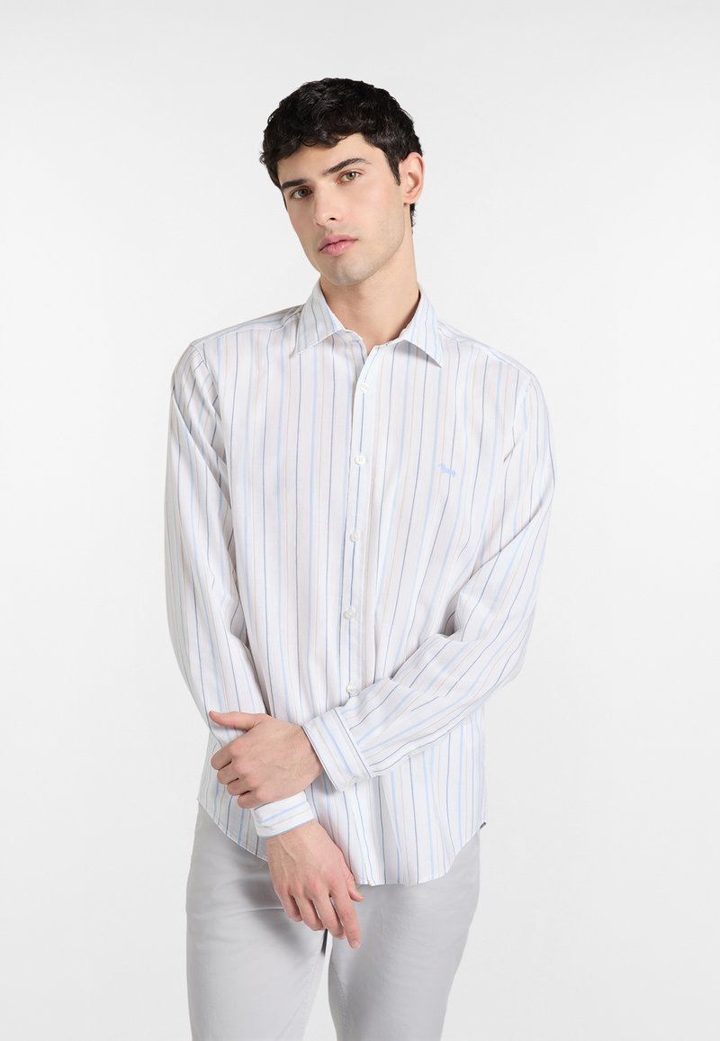 Jeune homme portant une chemise blanche à fines rayures verticales bleues et beiges, se tenant les bras croisés devant un fond blanc.