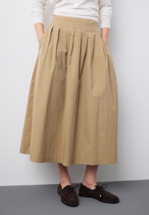 Gonna midi plissettata beige indossata con una maglia bianca a maniche lunghe e mocassini marrone scuro allacciati.