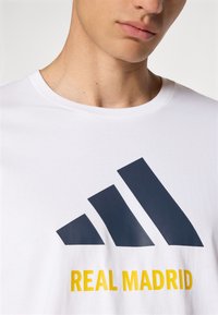 Hvid bomulds t-shirt med et stort navy blå grafik af tre diagonale striber og teksten "REAL MADRID" i gul.