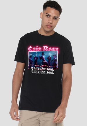Young man with short curly hair wears a black graphic t-shirt with neon text "Saia Boys" and silhouettes with the phrase "Ignite the soul."