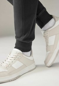 Jasnoszare sportowe sneakersy z białymi akcentami, materiałem o teksturowej powierzchni i gumową podeszwą, zestawione z czarnymi prążkowanymi spodniami dresowymi oraz szarymi skarpetami.