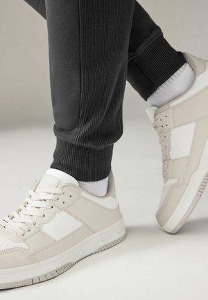 Light grey athletic sneakers with white accents, textured material, and a rubber sole, paired with black ribbed sweatpants and grey socks.