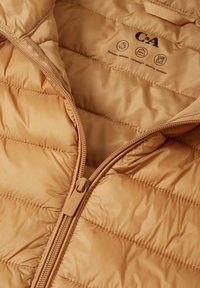 Chaqueta aislante beige con patrón acolchado, material ligero, cremallera color canela y etiquetas de cuidado que indican que está hecha de materiales reciclados.