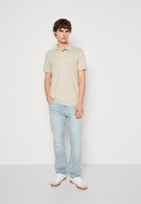 Πολυτελές πουκάμισο πόλο σε μπεζ απόχρωση με κοντά μανίκια, γιακά και λογότυπο; συνδυασμένο με ανοιχτό μπλε φθαρμένα τζιν και λευκά αθλητικά μποτάκια.