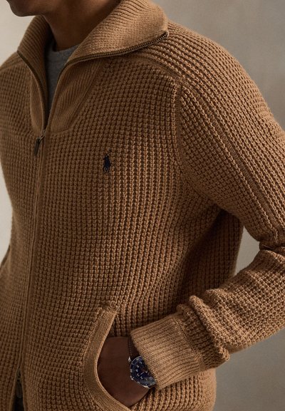 Bărbat purtând un pulover maro texturat cu fermoar și buzunare, cu mâna în buzunar și un ceas albastru la încheietură, pe un fundal neutru.