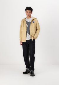Beige hettegenser med glidelås og snor, frontlomme og blå blomsterdesign på brystet, båret over en grå t-skjorte med grafikk og mørke jeans.