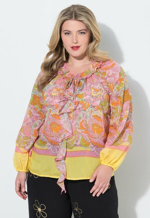 Kvinde med langt blondt hår iført en blomstret bluse i lyserød, orange og gul med flæser samt sorte bukser med guldbroderede blomster.