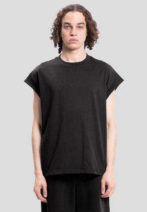 Persona con cabello rizado que lleva una camiseta negra sin mangas y holgada, de pie frente a un fondo liso de color gris claro.