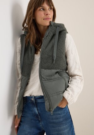 Gilet matelassé gris avec capuche et partie supérieure en polaire verte texturée, porté par-dessus un pull crème texturé. Un jeans bleu complète la tenue.