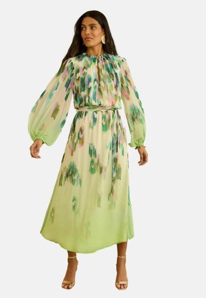 Frau trägt ein langärmeliges Midikleid mit abstraktem Muster in Pastellgrün, Lila und Blau, tailliert und kombiniert mit goldenen High-Heeled Sandalen.