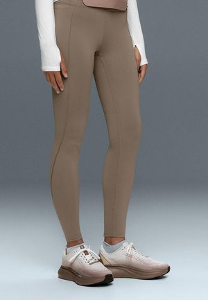Personne portant des leggings taupe ajustés, un haut blanc à manches longues et des chaussures de sport blanches avec des semelles beiges, debout sur un sol gris clair.