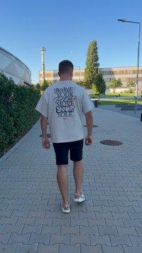 Fény bézs, oversized póló, fekete és piros felirattal a hátsó részén. Fekete rövidnadrággal és fehér tornacipővel párosítva, hátulról nézve.