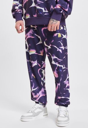 Lilla joggebukser med et fargerikt abstrakt mønster, elastiske mansjetter og en logodetalj. Kombinert med hvite og grå joggesko.