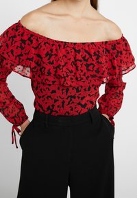 Blouse rouge à épaules dénudées avec motif floral noir, détail à volants superposés et manches longues avec poignets élastiques, associée à un pantalon noir.