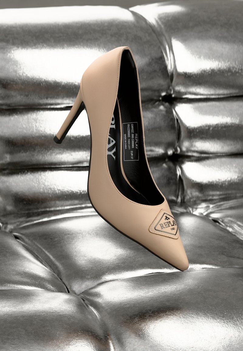 Chaussure à talon haut beige avec embout pointu et un accent noir. Fabriquée en cuir lisse, elle présente un patch logo sur le dessus, posée sur une surface argentée.