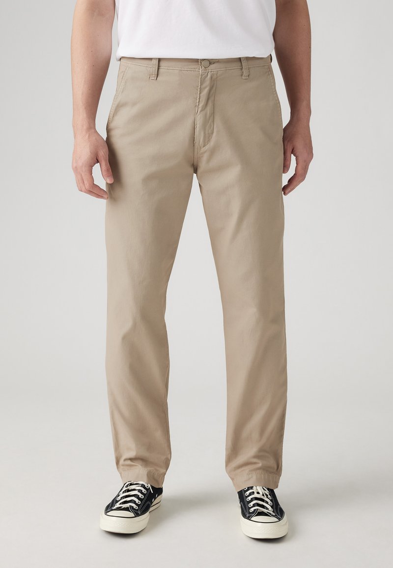 Chinos en coton beige avec une coupe droite, dotés de poches avant et d'une fermeture éclair, associés à des baskets noir et blanc.