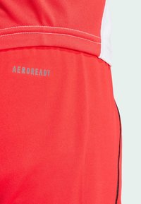 Punaiset urheilushortsit, joissa on hengittävä kangas ja kohokuvioitu "AEROREADY" -logo. Sivusaumojen ääriviivat mustana, sileä rakenne.