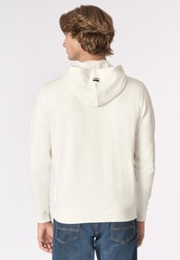 Uomo che indossa un hoodie bianco con una piccola striscia rossa, bianca e blu sul cappuccio, abbinato a jeans blu, visto da dietro.