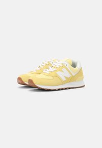 Een paar gele en witte New Balance sneakers met witte veters en gumrubberen zolen op een witte achtergrond.